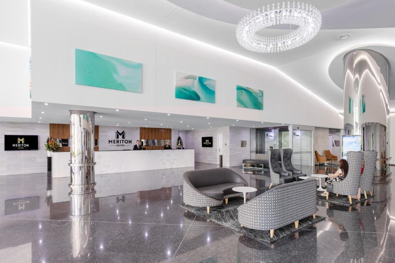 Meriton Suites Southport Gold Coast Buitenkant foto The photo shows a modern hotel lobby with a sleek and stylish design. Key features include:

- A reception desk at the back with the name "Meriton" displayed prominently.
- Elegant lighting, including a chandelier hanging from the ceiling.
- The floo