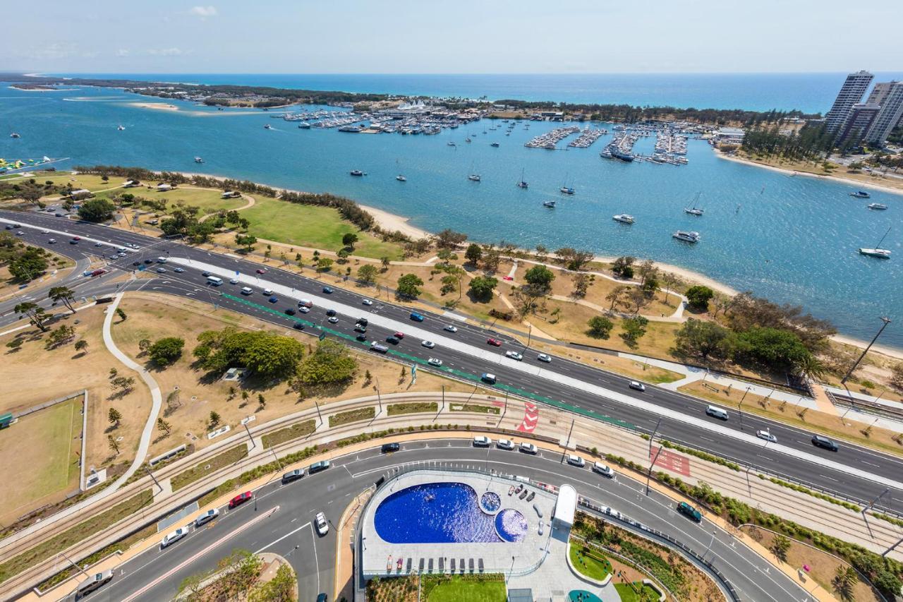 Meriton Suites Southport Gold Coast Buitenkant foto The photo shows an aerial view of a coastal area. In the foreground, there is a roundabout with a large blue pool and landscaped green spaces. Surrounding the roundabout is a roadway with vehicles. Beyond the road, there are patches of grass and tree