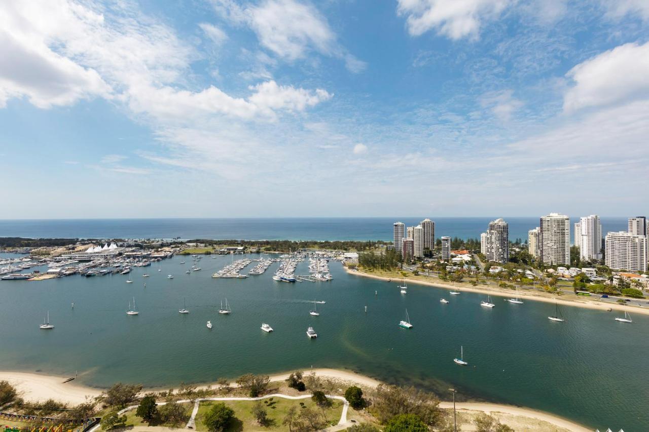 Meriton Suites Southport Gold Coast Buitenkant foto The photo depicts a scenic coastal view featuring a marina with several boats docked. In the foreground, there is a river or inlet with a sandy beach area. The background showcases a stretch of blue ocean and a clear sky dotted with a few fluffy clou