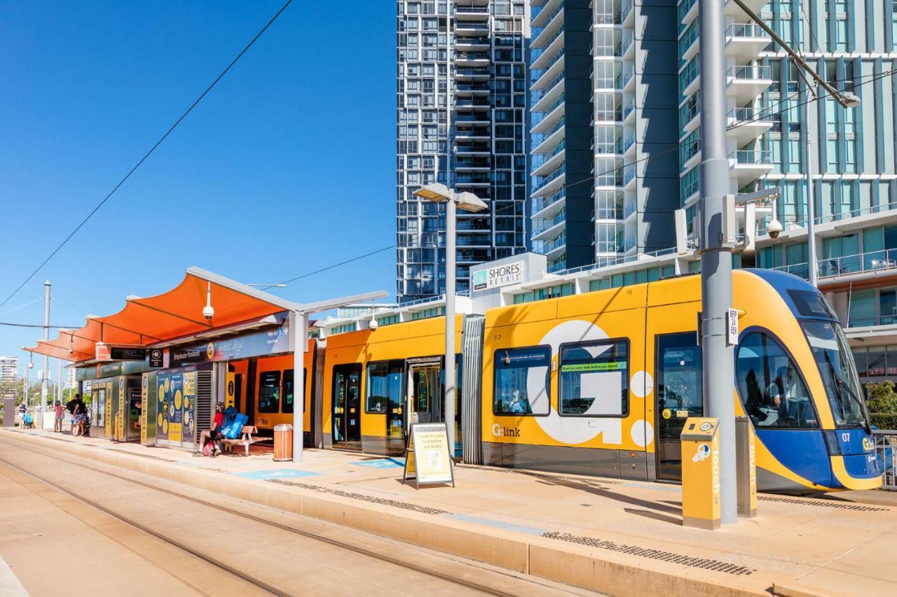 Meriton Suites Southport Gold Coast Buitenkant foto The photo shows a modern tram station with a brightly colored yellow tram. The station features an orange canopy, providing shade for passengers. There are people seated nearby, and tall buildings are visible in the background against a clear blue sk