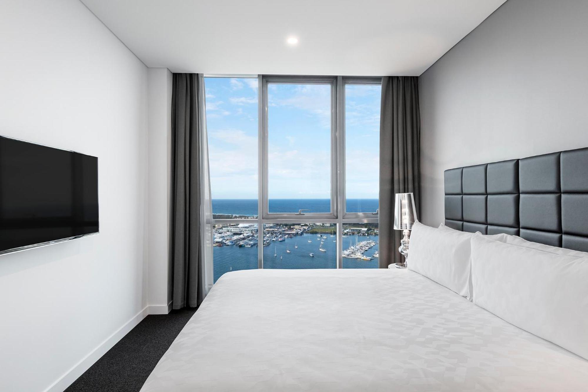 Meriton Suites Southport Gold Coast Buitenkant foto The photo shows a modern hotel room featuring a large bed with white linens and dark accent pillows. To the left, there is a flat-screen TV mounted on the wall. The room has a large window with two curtains that allows natural light to enter and offe